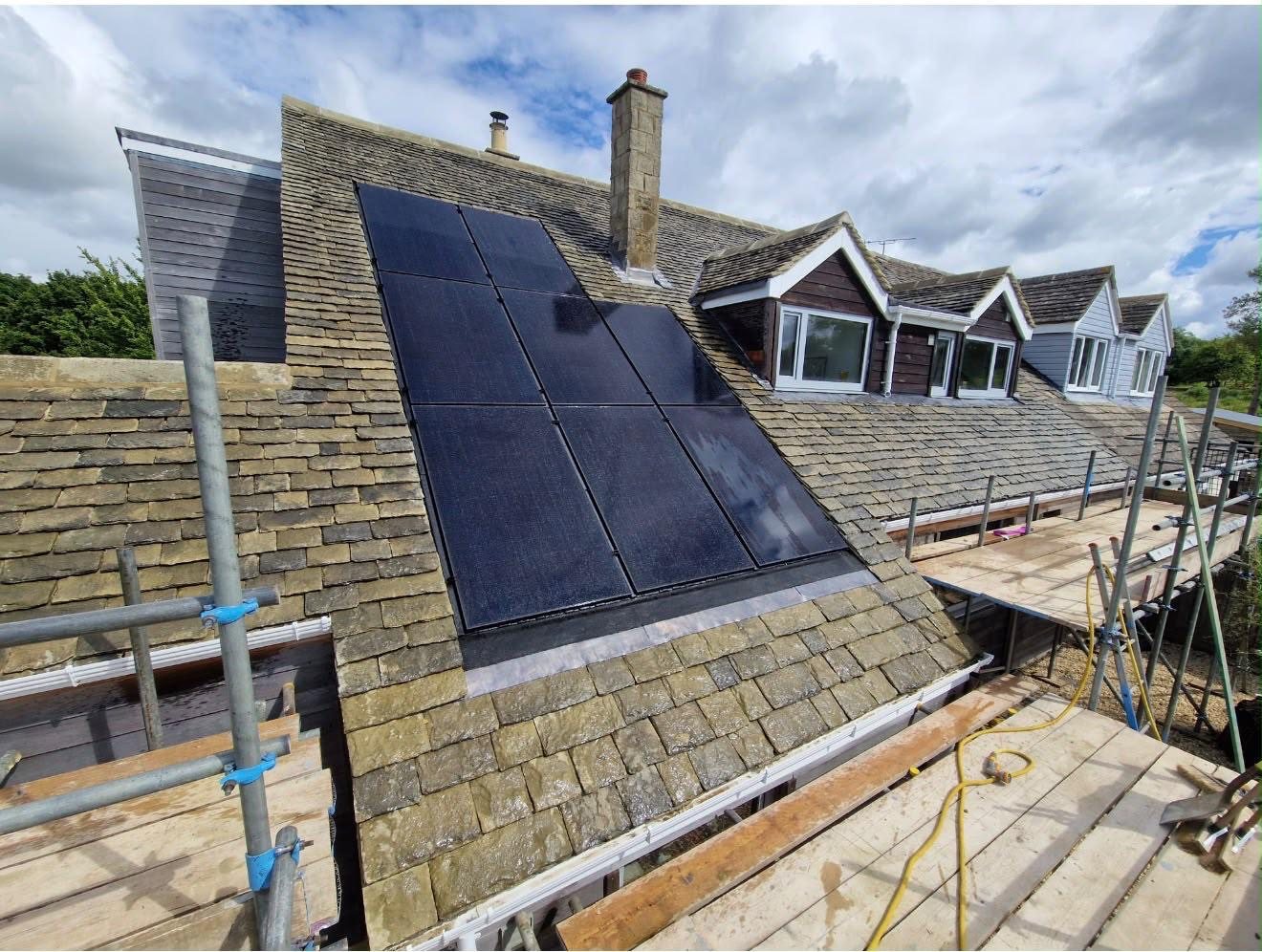 Black in-roof solar panels on a stone-slate roof with dormer windows, scaffold in the foreground