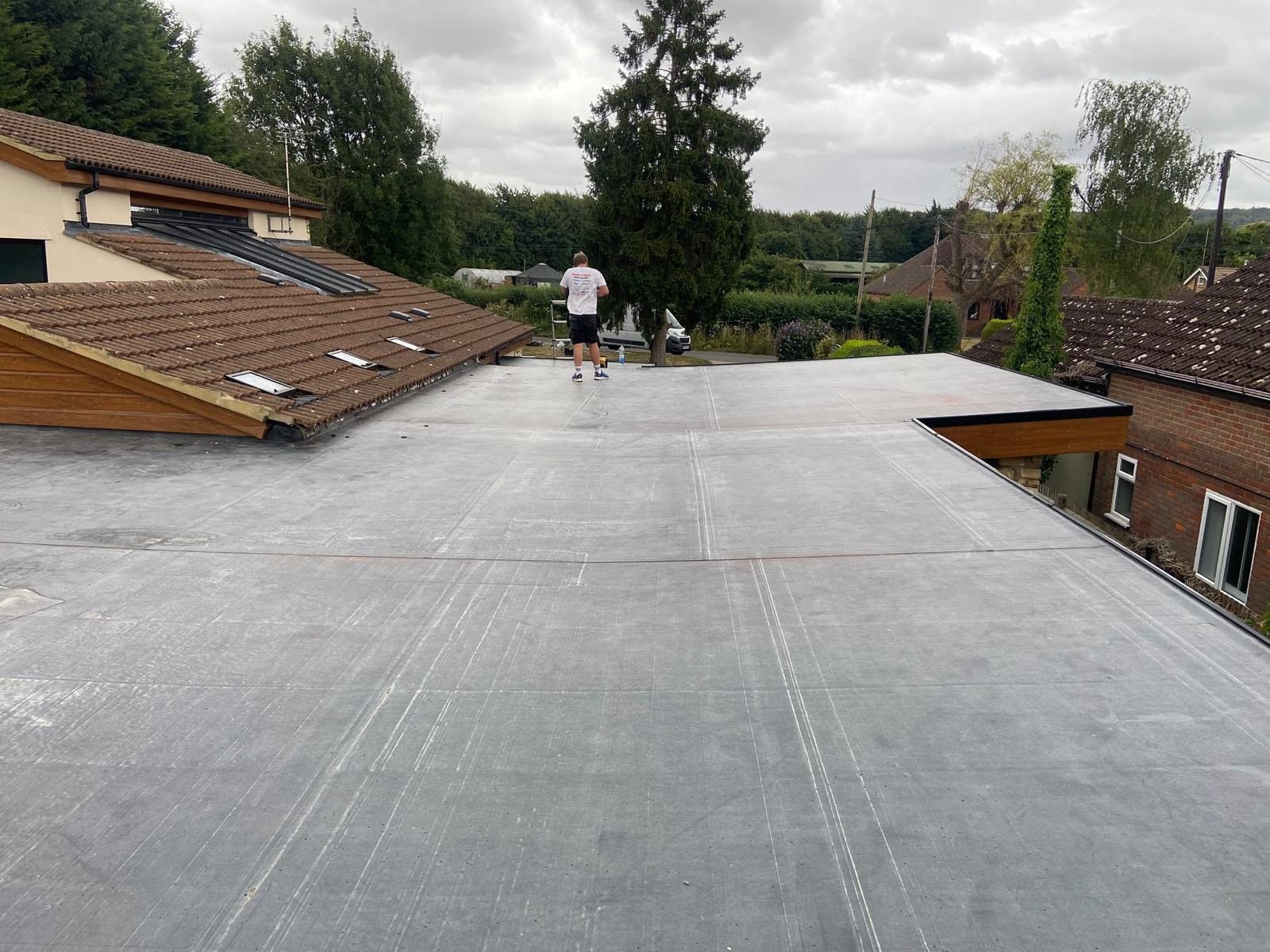Large new grey EPDM rubber flat roof with a roofer standing on it, trees behind, overcast sky