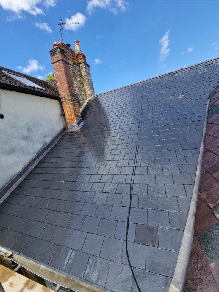 Natural slate pitched roof with a brick chimney and lead flashing, under a blue sky